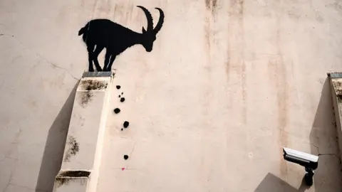 PA Media The black silhouette of a goat stands on the external structural support of a building with falling painted black rocks, and a CCTV camera points in its direction.